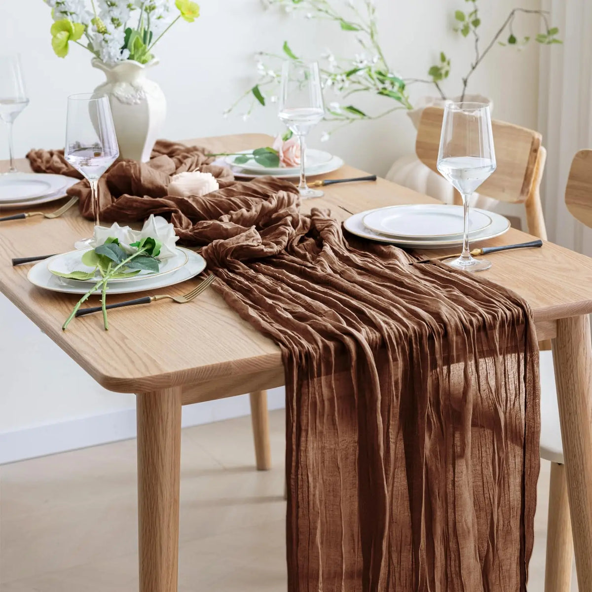 A brown cheesecloth table runner draped on a wooden table set for a meal with plates and glassware.