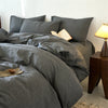 A gray yarn-dyed duvet cover is displayed on a bed with pillows and a book, next to a wooden lamp.