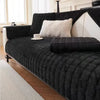 A black quilted sofa protection cover with white pillows and a light-colored rug in a bright room.