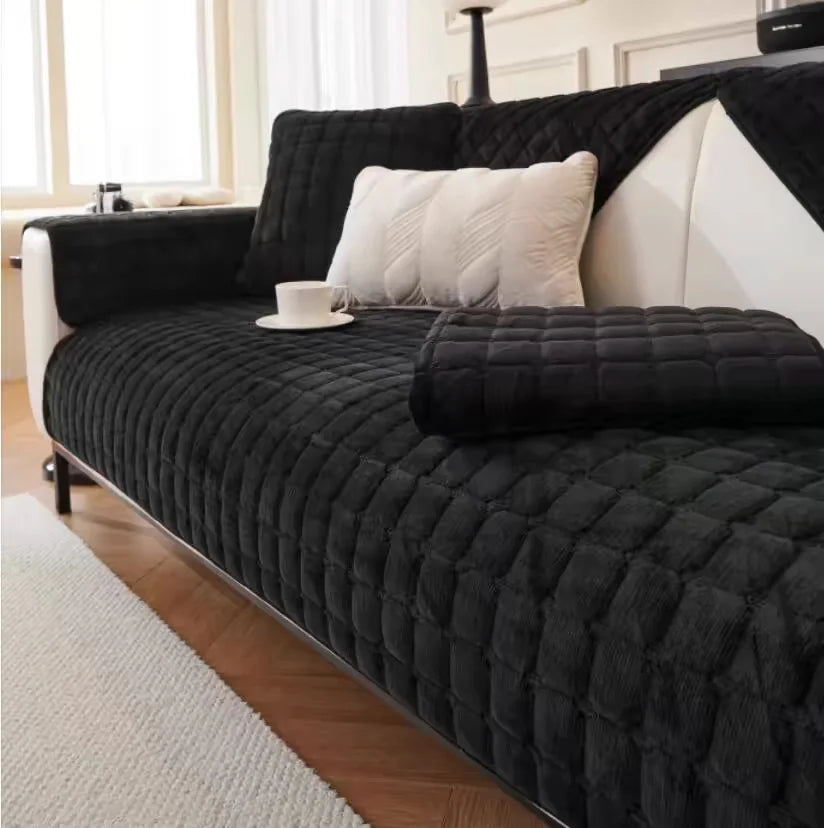 A black quilted sofa protection cover with white pillows and a light-colored rug in a bright room.