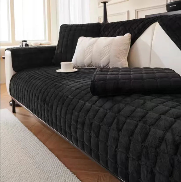 A black quilted sofa protection cover with white pillows and a light-colored rug in a bright room.