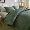A quilted olive green lightweight queen quilt spread on a bed with white linens and light wood furniture.