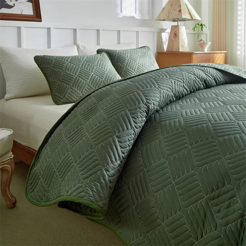 A quilted olive green lightweight queen quilt spread on a bed with white linens and light wood furniture.