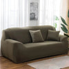 A modern, taupe stretch sofa cover placed in a bright living room with sheer curtains and a potted plant.