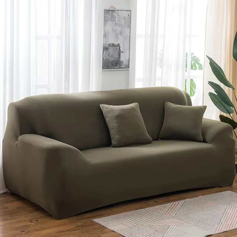 A modern, taupe stretch sofa cover placed in a bright living room with sheer curtains and a potted plant.