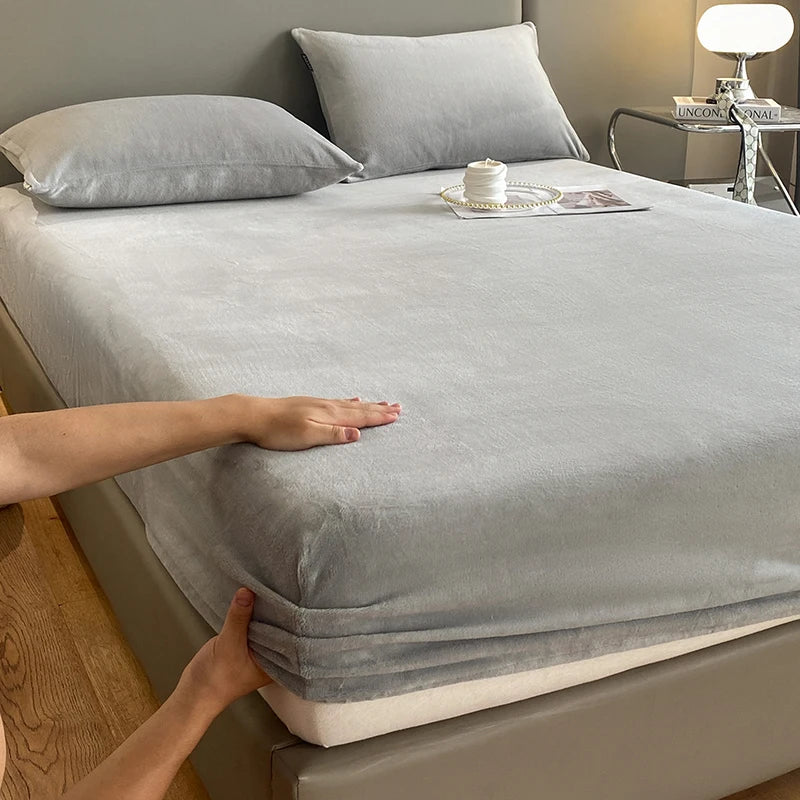 A hand is touching a gray luxury velvet bedding set on a bed, with pillows and a lamp visible in the background.