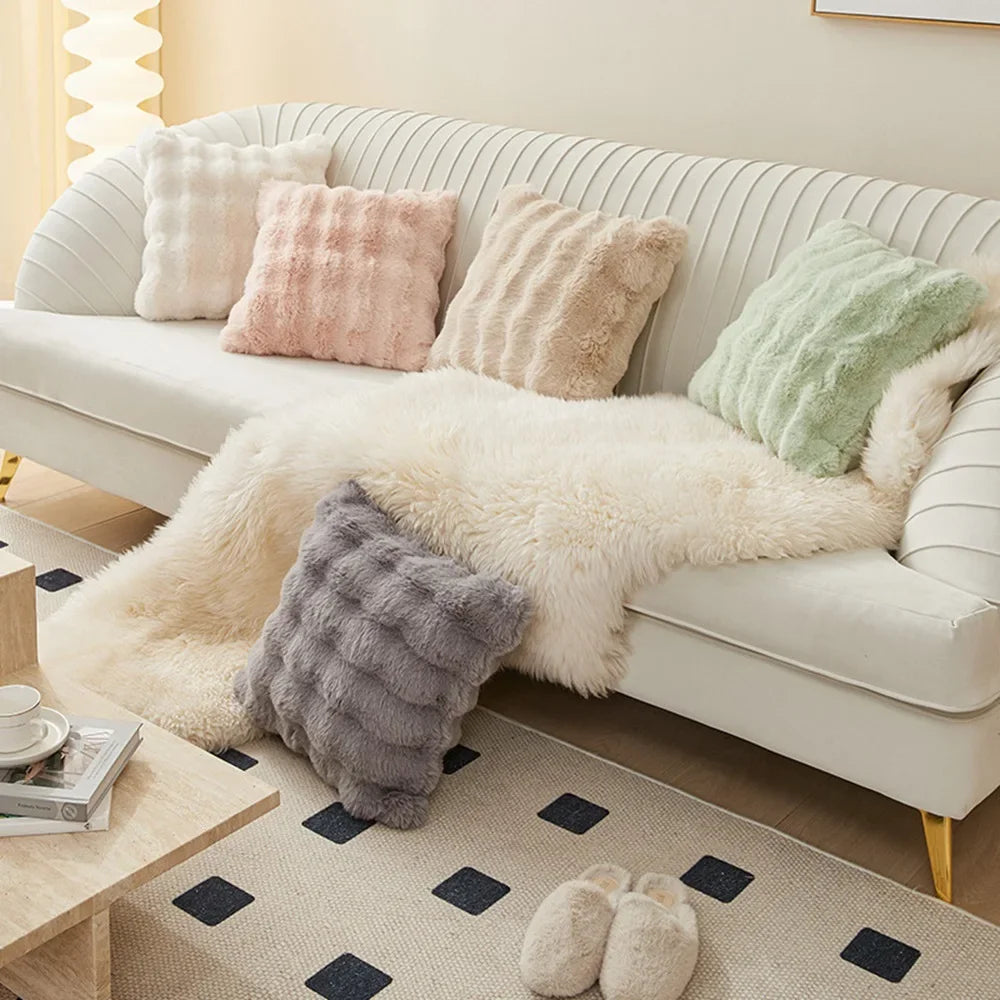A white sofa adorned with various fluffy and textured cushion covers in pastel shades of pink, cream, and green.