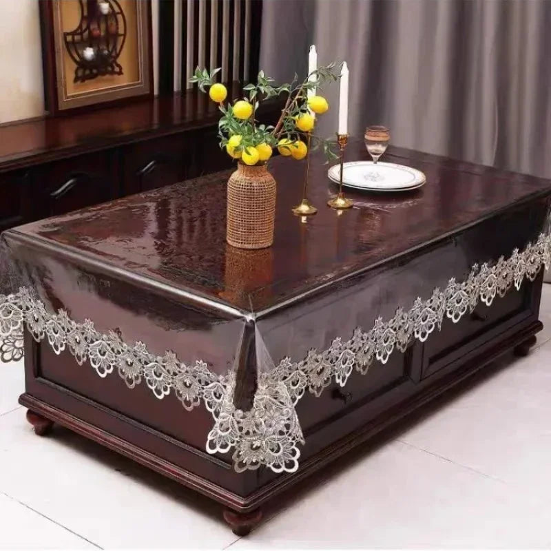 A dark wood table covered with a lace embroidered PVC tablecloth featuring yellow fruit and candles.