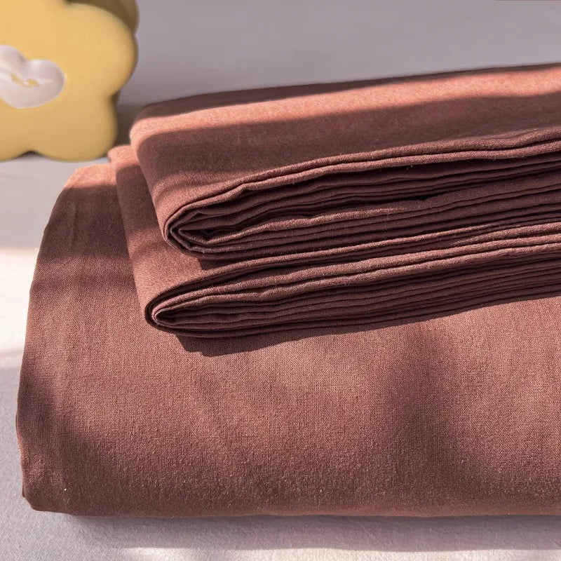A stack of brown organic cotton bedding, including a flat sheet and pillowcases, is displayed on a light surface.