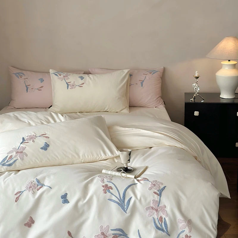 A white duvet cover set with embroidered pink and blue orchids and a lace trim on a bed.