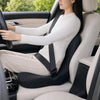 A woman is sitting in a car seat with a white top and light pants, using an ergonomic memory foam cushion.