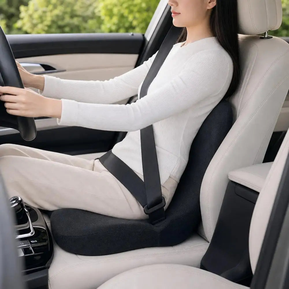 A woman is sitting in a car seat with a white top and light pants, using an ergonomic memory foam cushion.