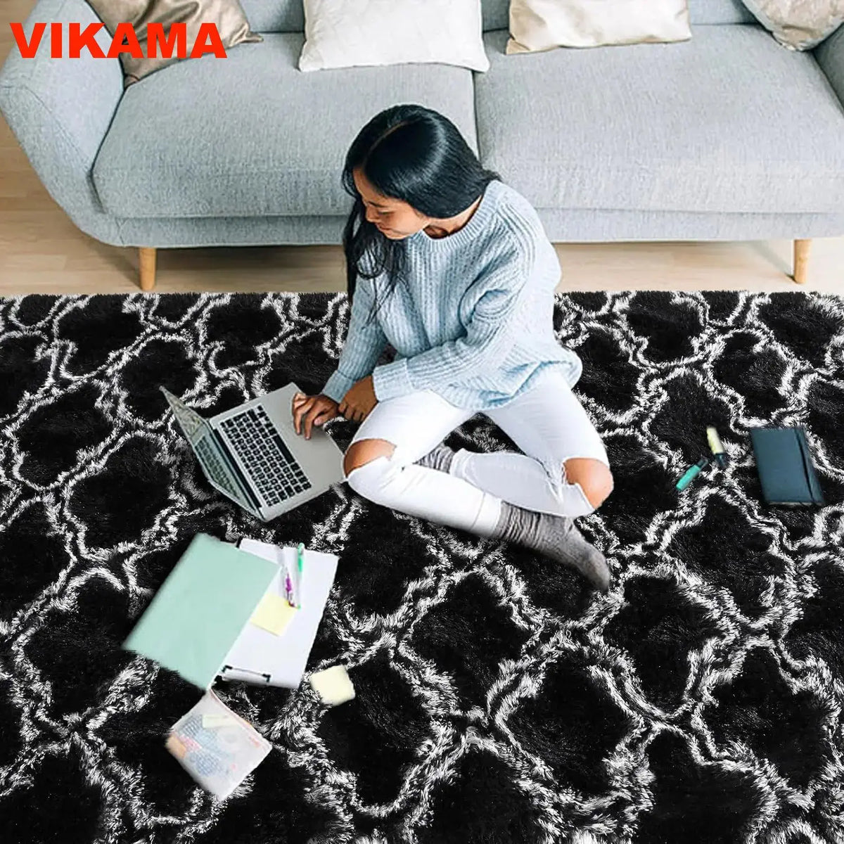 A person sits on a black geometric plush area rug in a living room, working on a laptop.