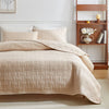 A beige quilted cotton bedspread and pillows displayed on a bed with a wooden shelf in the background.