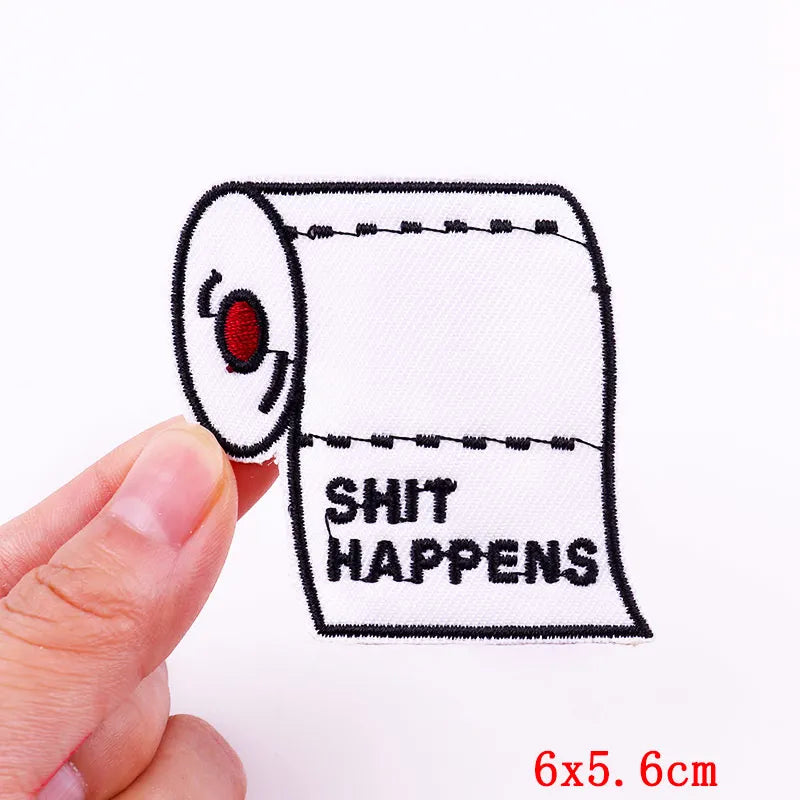 A white embroidered patch with the text SHIT HAPPENS on a fabric roll being held by a hand.