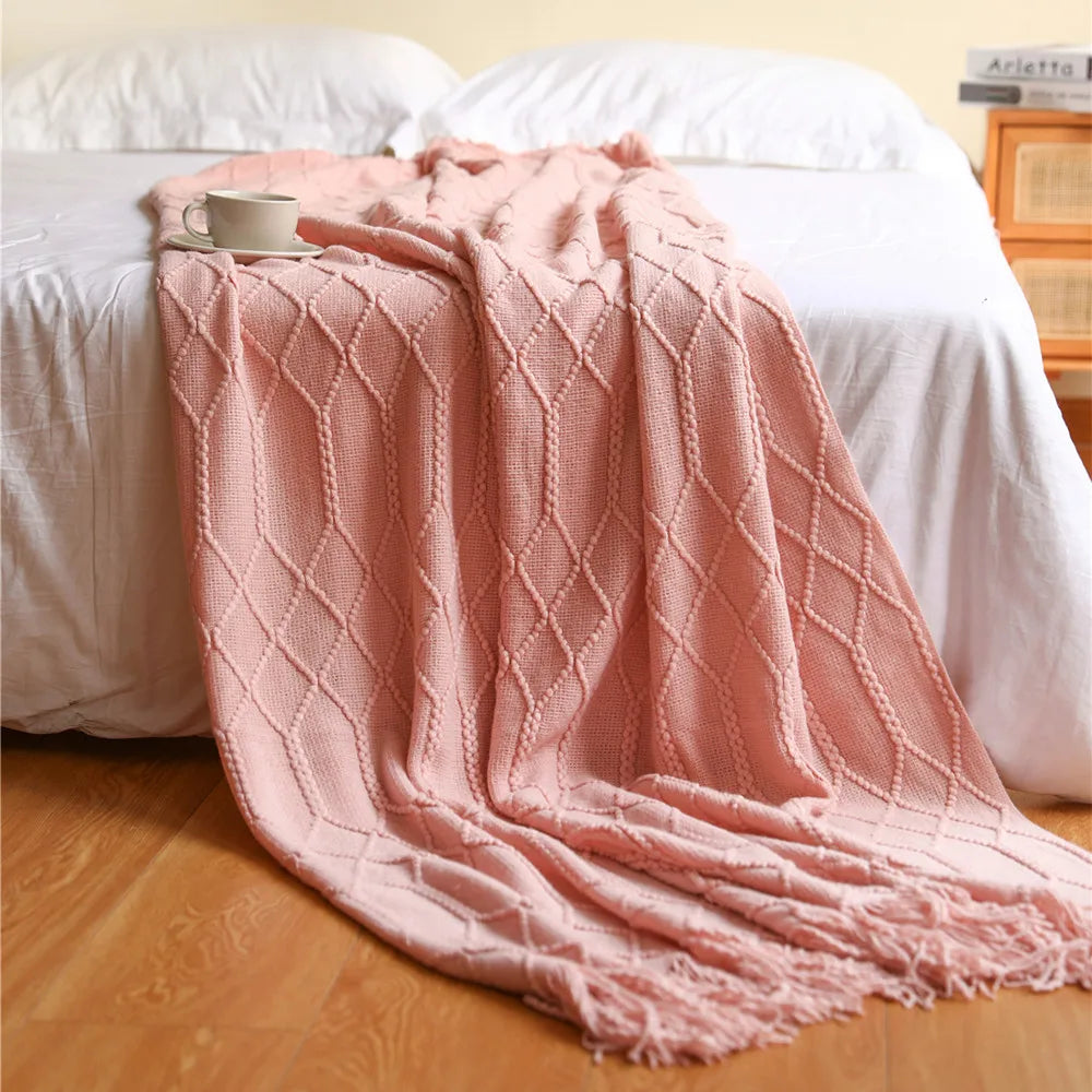 A soft pink knitted tassel throw blanket draped over a white bed with white linens.