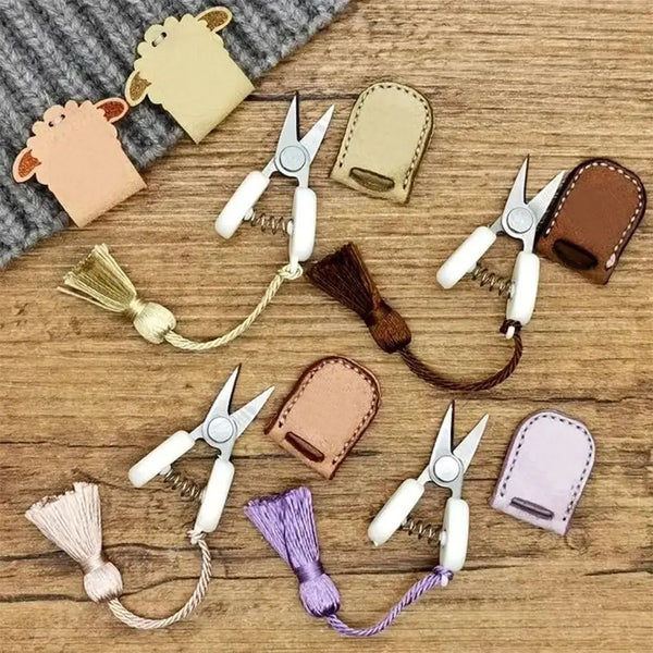 A collection of mini precision sewing scissors in various colors and styles with protective leather covers on a wooden surfac