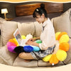 A woman is sitting on a sofa surrounded by yellow and orange plush cushions, looking at a laptop.