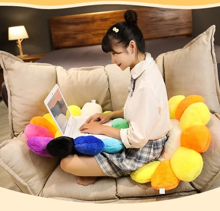 A woman is sitting on a sofa surrounded by yellow and orange plush cushions, looking at a laptop.