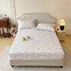 A white floral printed cotton fitted sheet is displayed on a bed with a light upholstered headboard and bedding.