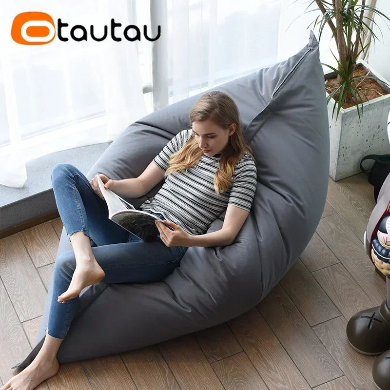 A young woman relaxing in a gray outdoor bean bag cover on a wooden floor while reading a book.