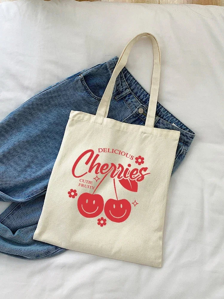 A cream canvas shopping bag featuring the text Delicious Cherries and two smiling cherry illustrations, resting on a denim sh