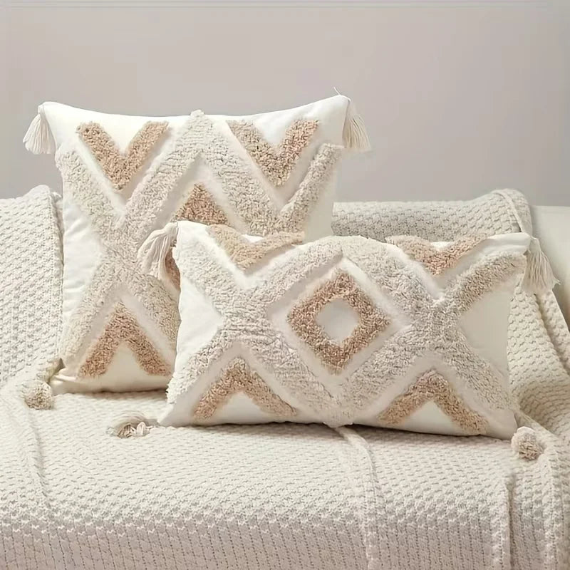 A white plush pillow cover with beige geometric embroidery and tassel details resting on a cream textured fabric.