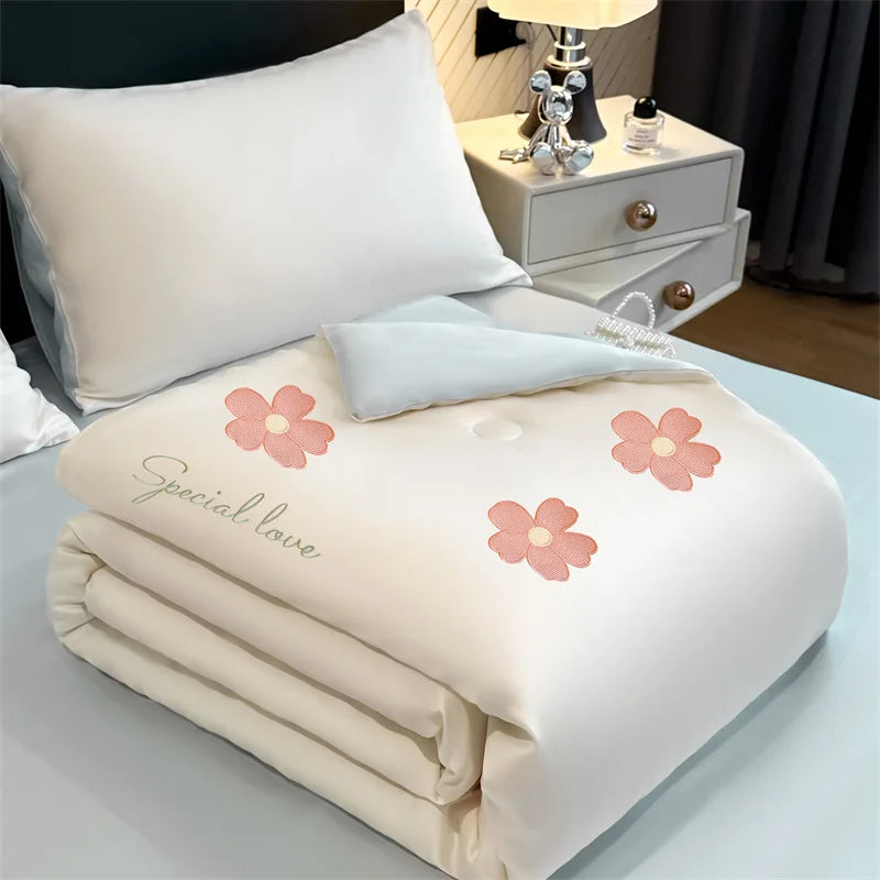 A white embroidered cooling summer quilt with pink floral designs is displayed on a light blue surface.