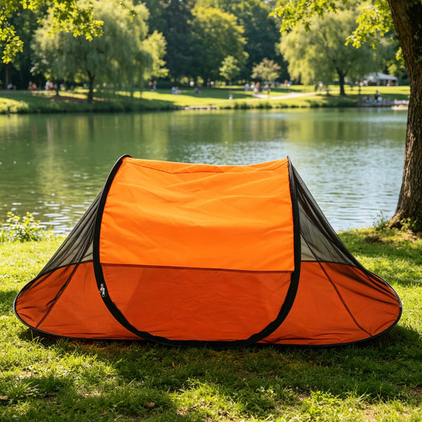 An orange portable pop-up mosquito net shelter laid out on green grass beside a pond.