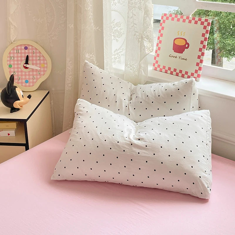 A pink bed features a white duvet cover and pillows with black polka dots, next to a decorative clock and a framed sign.