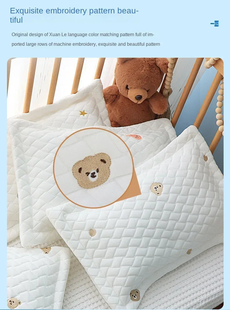 A white quilted pillow with a bear design is shown next to a brown teddy bear on a wooden crib railing.