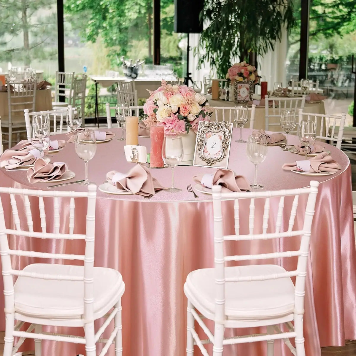 A round dining table set for an elegant event featuring pink satin tablecloths, white chairs, and floral centerpieces.