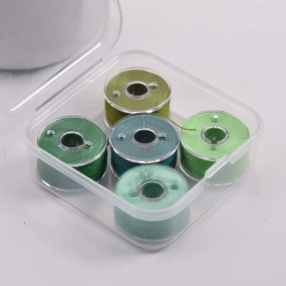 A clear plastic container holds several spools of prewound sewing machine bobbin threads in various shades of green.