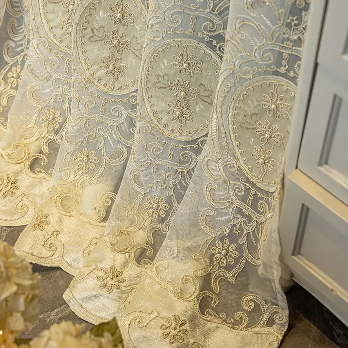Ivory embroidered beaded sheer curtain with punched-hole design hanging next to a white cabinet.