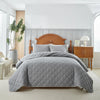A gray quilted bedspread set is displayed on a bed with a wooden headboard and bedside table in a bright bedroom.