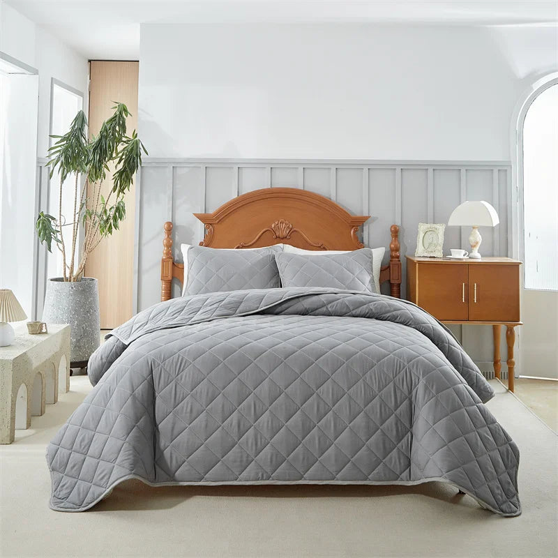A gray quilted bedspread set is displayed on a bed with a wooden headboard and bedside table in a bright bedroom.