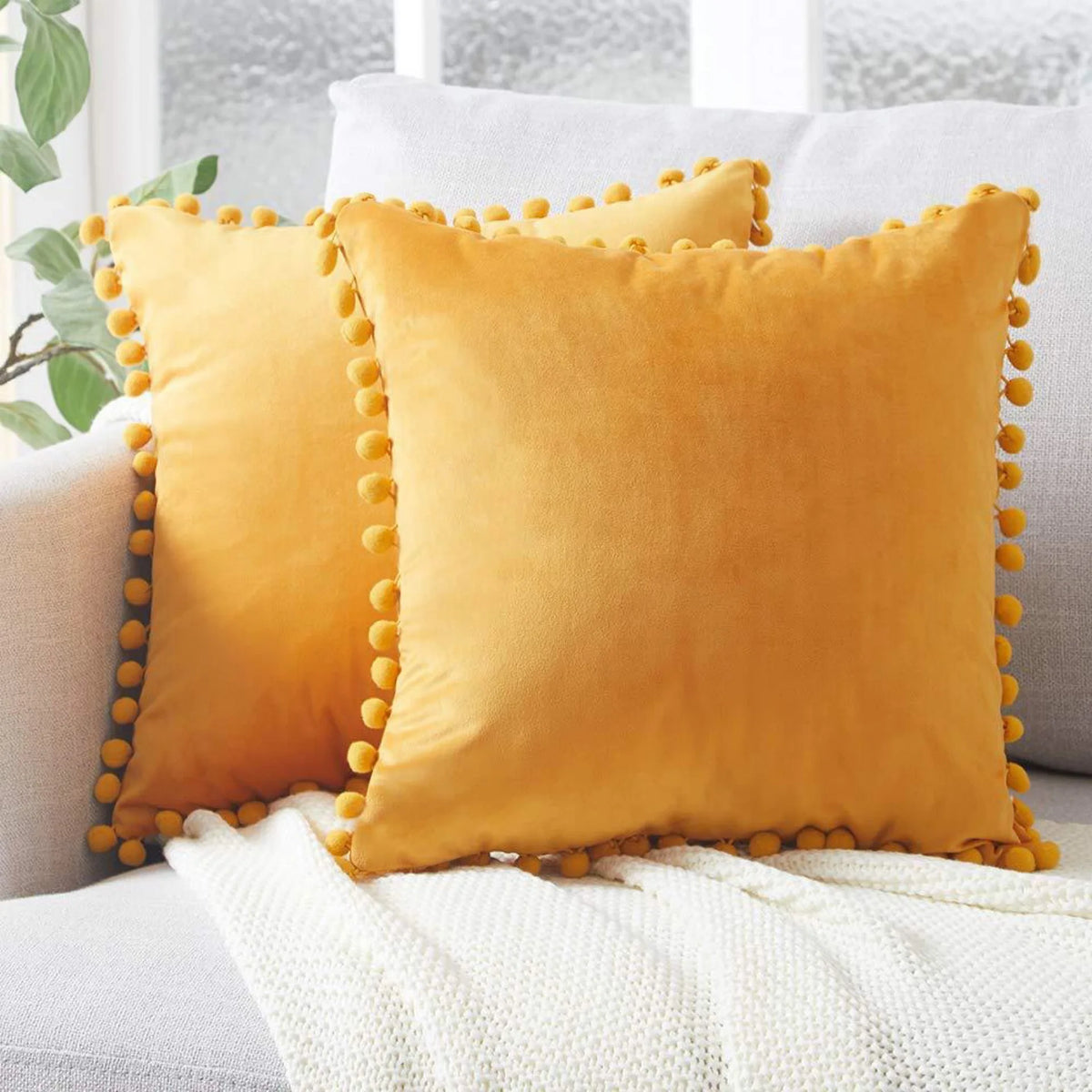 A square mustard yellow velvet cushion with gold trim resting on a white knitted blanket.