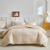 A beige lightweight queen quilt bedding set displayed on a bed with white paneled walls and a wooden nightstand.