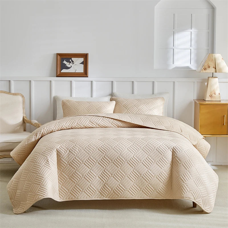 A beige lightweight queen quilt bedding set displayed on a bed with white paneled walls and a wooden nightstand.