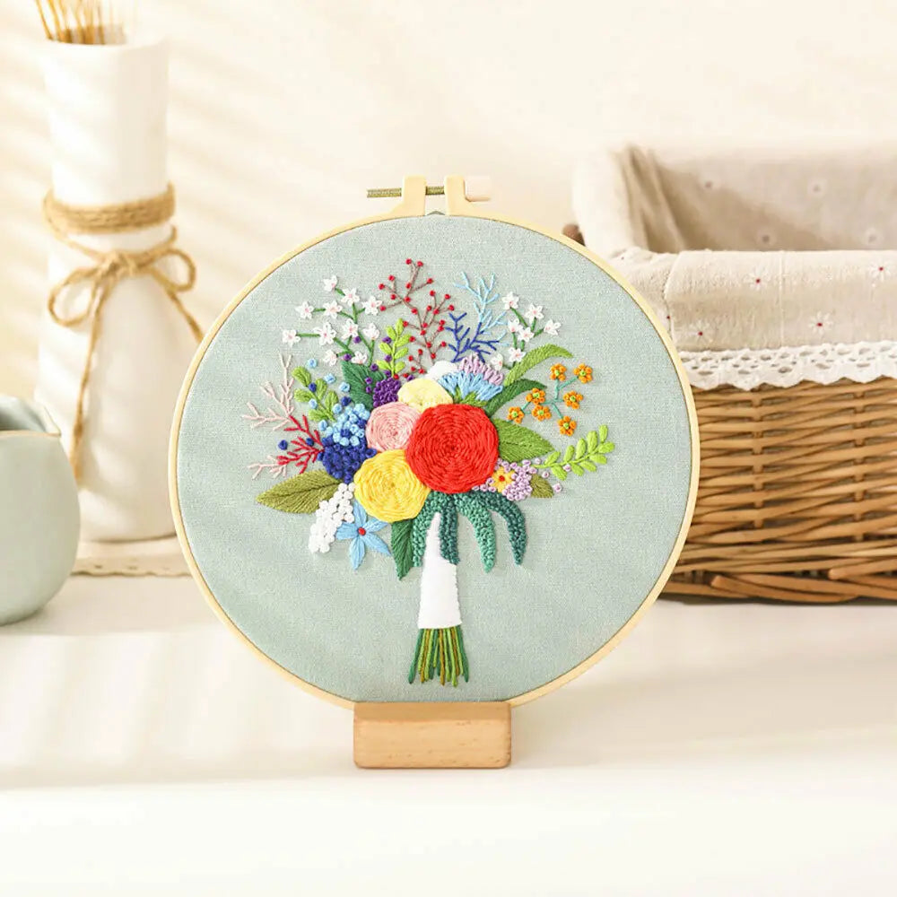A circular embroidery hoop featuring a colorful floral design with various flowers and greenery on a light teal fabric.