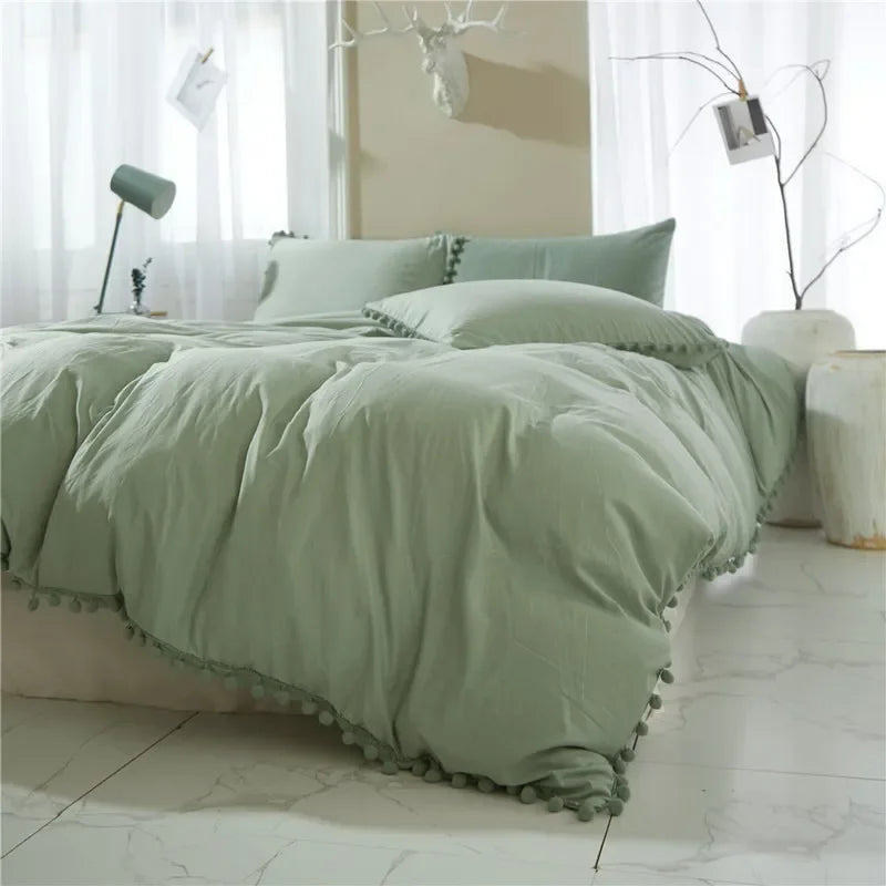A light sage green duvet cover set with pom-pom trim displayed on a bed in a bright room.