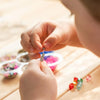 A child is using a stainless steel beading needle to thread beads onto a string in a craft setting.