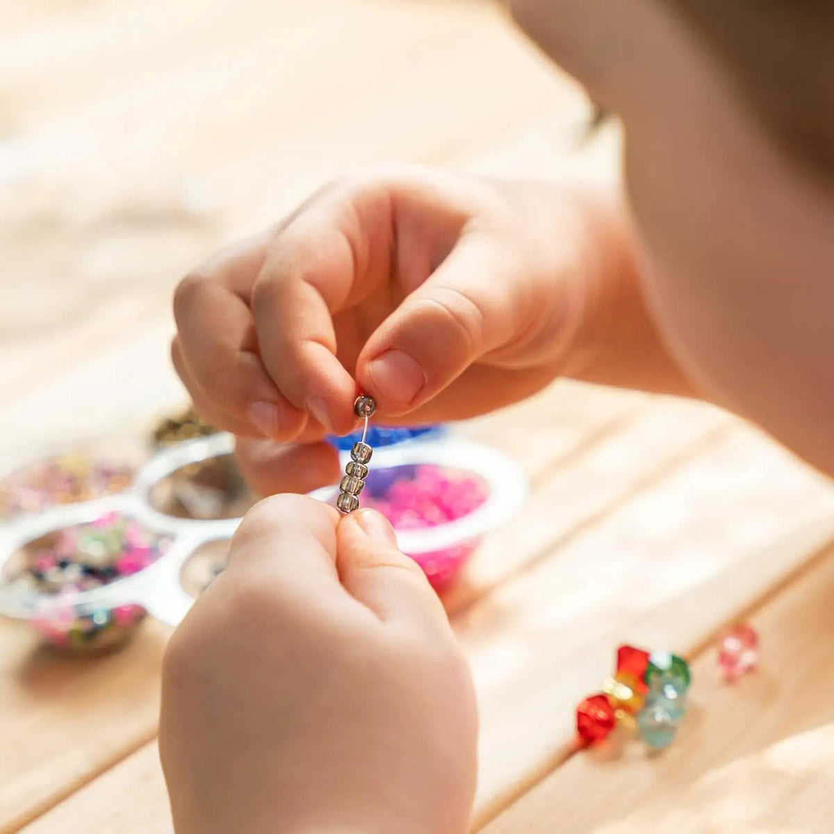 A child is using a stainless steel beading needle to thread beads onto a string in a craft setting.