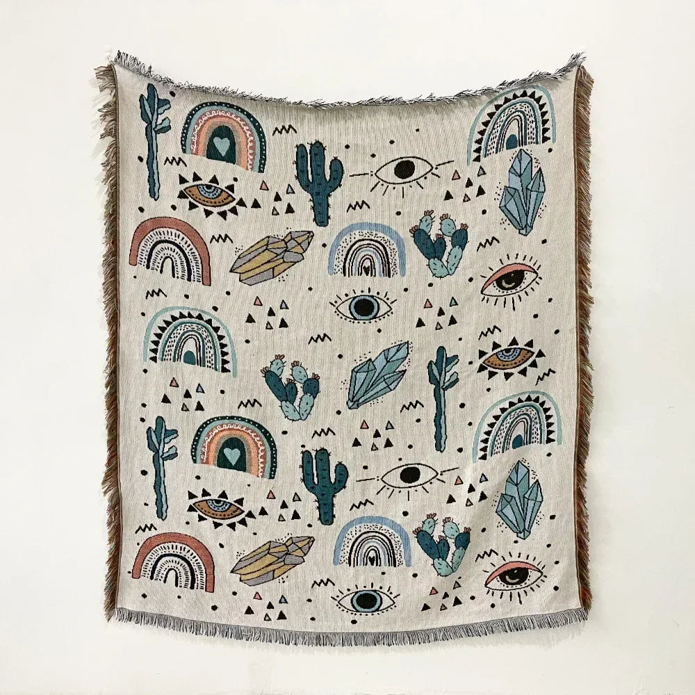 A throw blanket featuring a geometric pattern of cacti, eyes, and rainbows on a light fabric with fringed edges.