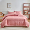 A pink quilted cotton bedspread set with matching pillows on a bed in a bright room.