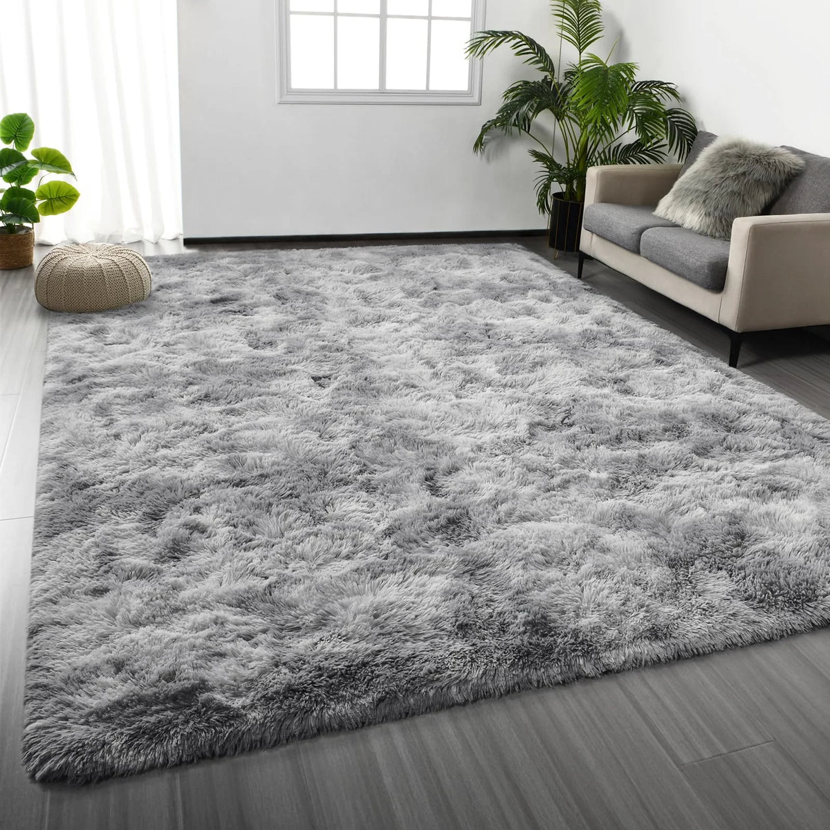 A gray shaggy rug is placed on a wooden floor in a bright room with a sofa and a large potted plant.