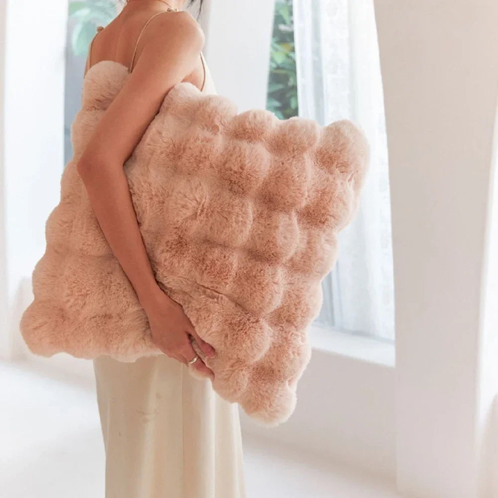 A person is holding a large, fluffy, light pink faux fur cushion cover against a bright window.