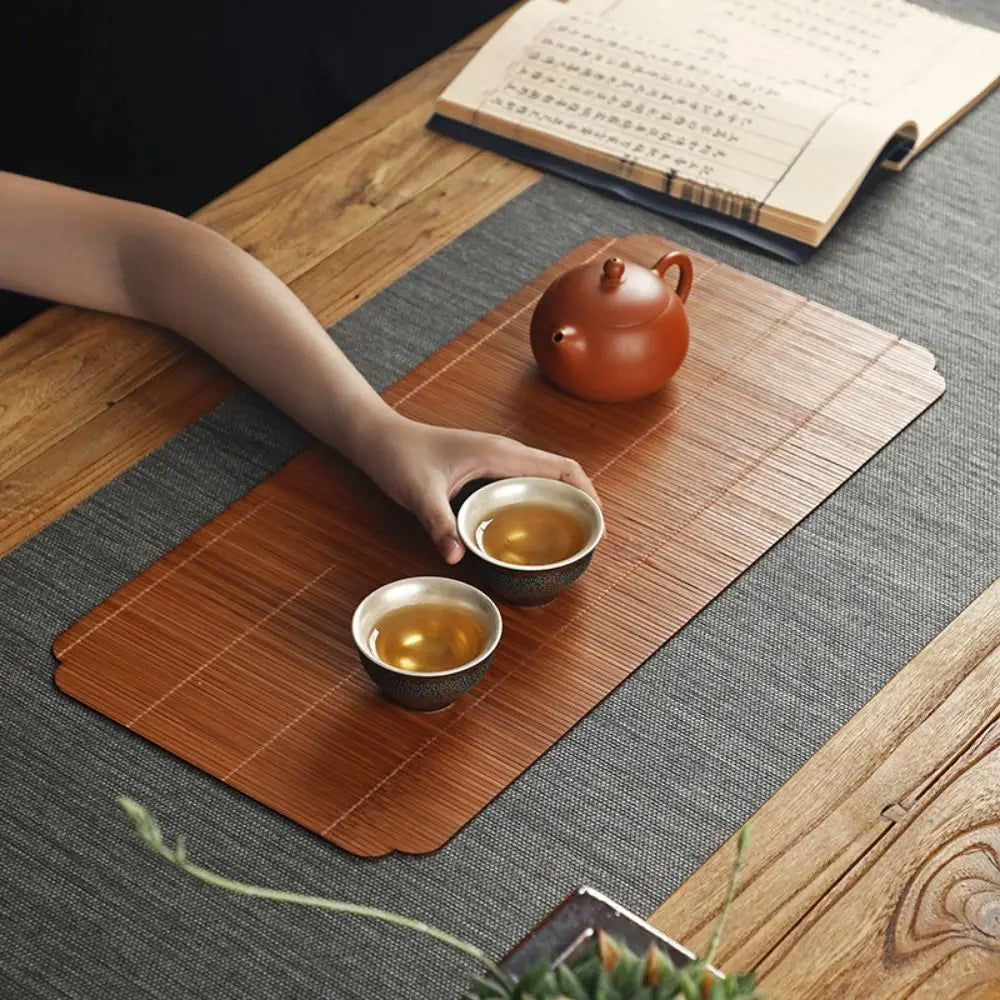 A hand-woven bamboo tea mat displaying a terracotta teapot and two small cups on a wooden table.