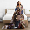 A woman is relaxing on a bed with a large throw blanket featuring various women's faces and text.