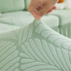 A hand touches a textured, sage green jacquard elastic sofa cover with a leaf pattern.